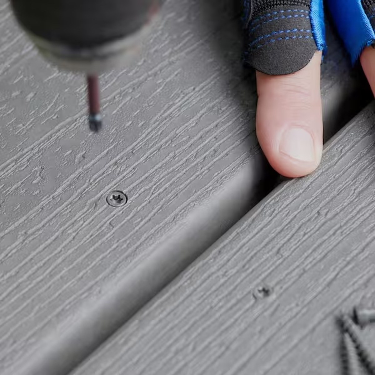 Close-up of a gloved hand steadying a grey wood-grain composite deck board while a driver drives a face screw through the surface, with an already-driven countersunk screw head sitting in its dish in the board beside it